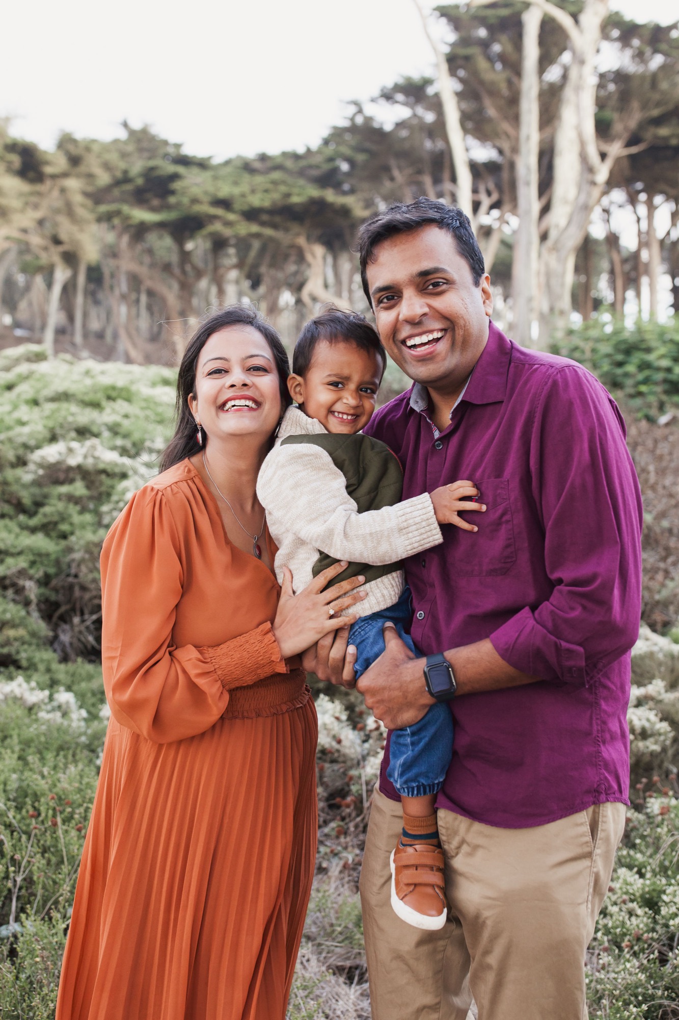 A family of three beams at the camera with genuine joy, framed by cypress trees and coastal vegetation under soft daylight