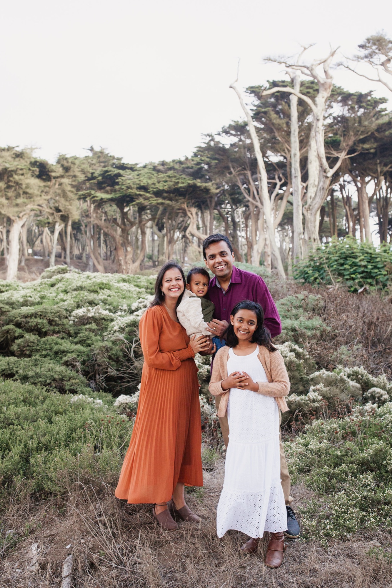 A family of four smiles warmly amid coastal sage scrub with majestic Monterey cypress trees towering behind them under soft overcast light