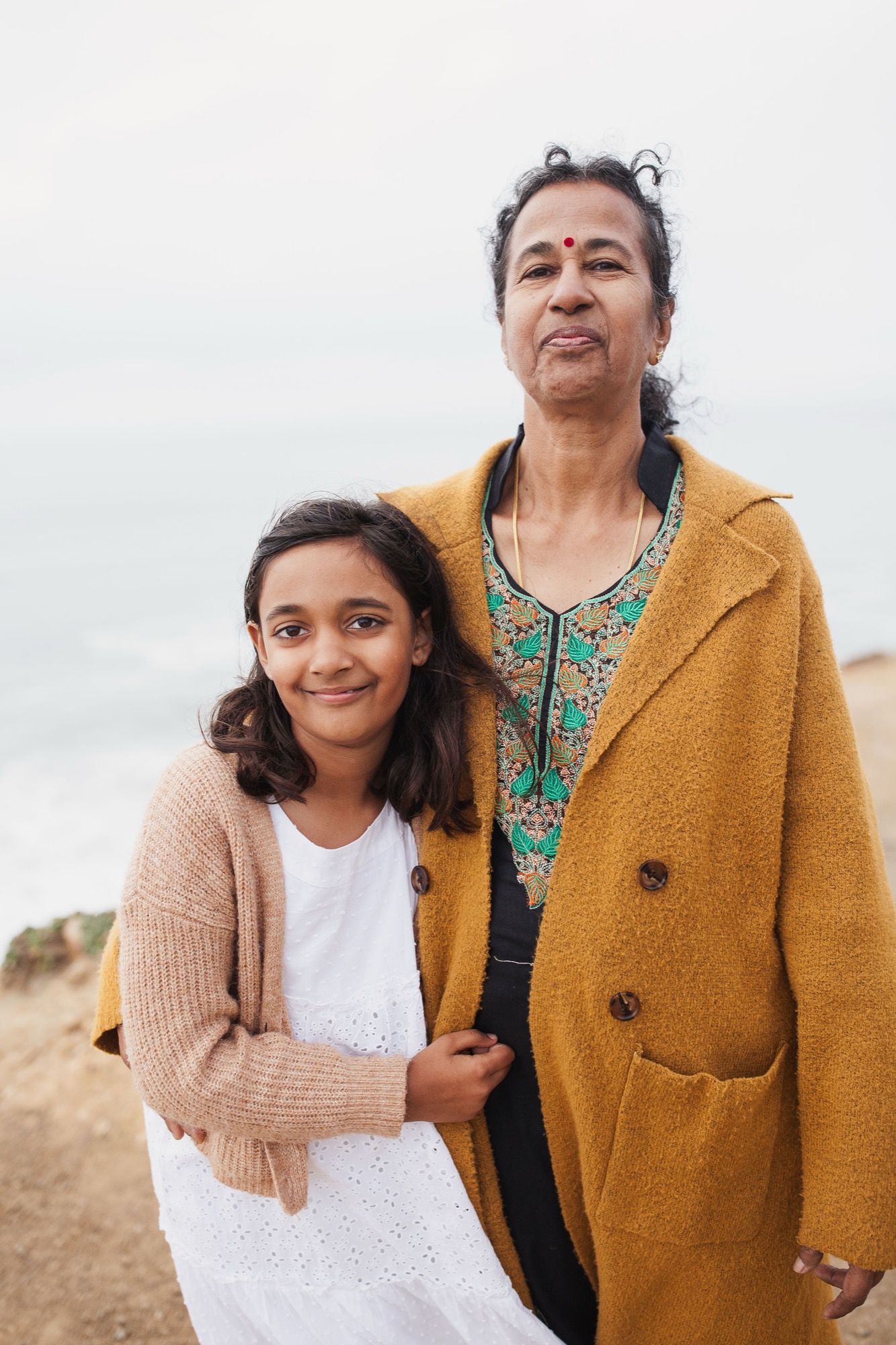 Woman and girl stand together on a sandy beach, the girl smiling warmly while the woman gazes upward, both wearing warm-toned cardigans against an overcast sky