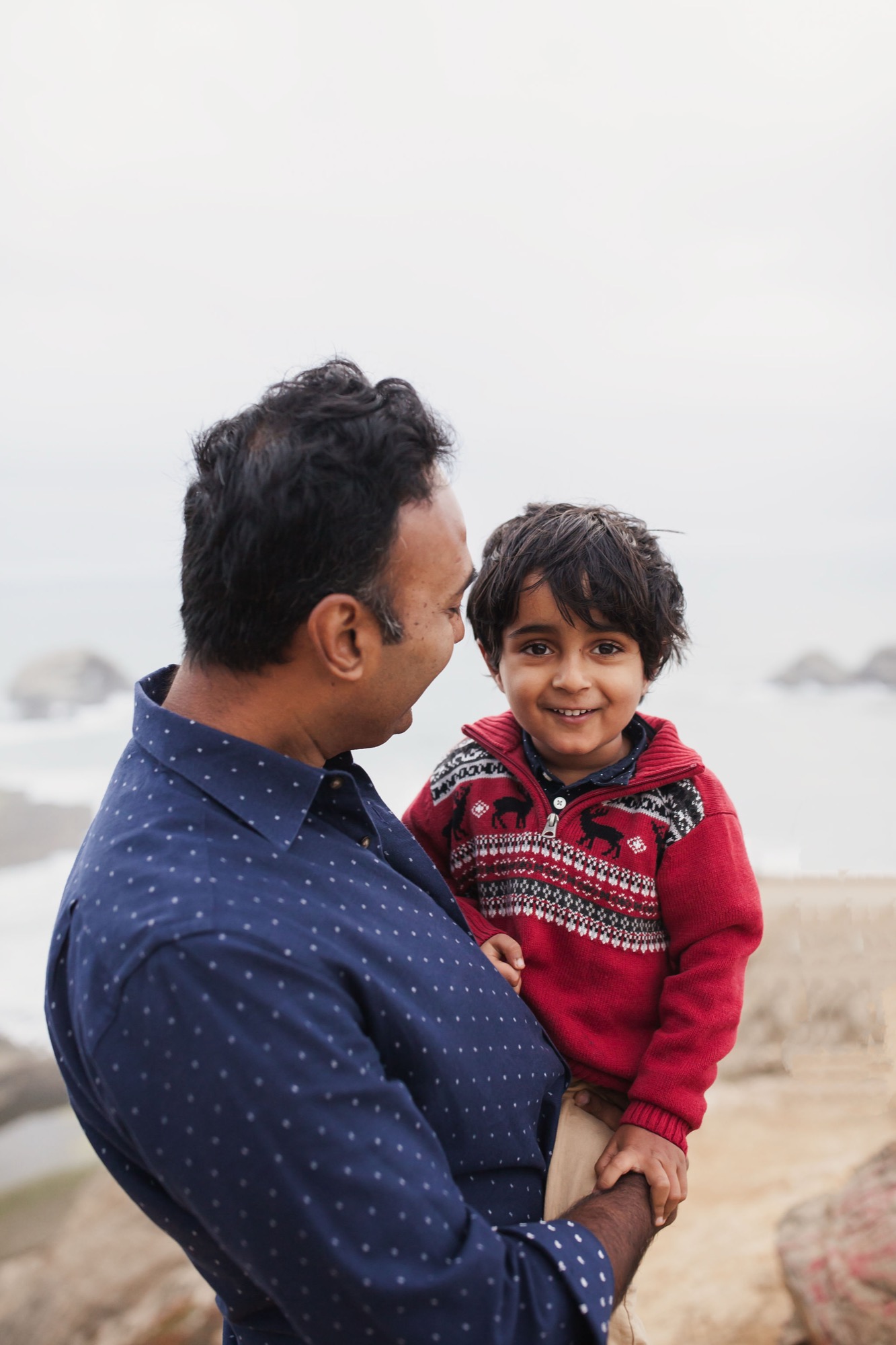 A father holds his young son on a misty beach, both smiling warmly as ocean waves blur softly behind them