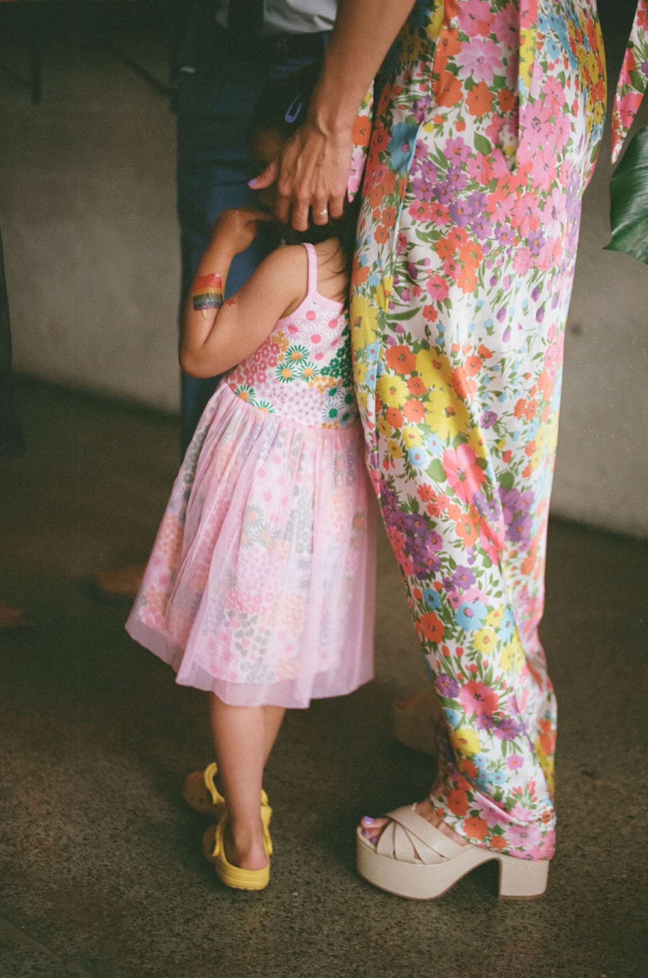 A small child in a pink floral dress holds an adult's hand, their yellow shoes visible against the concrete floor