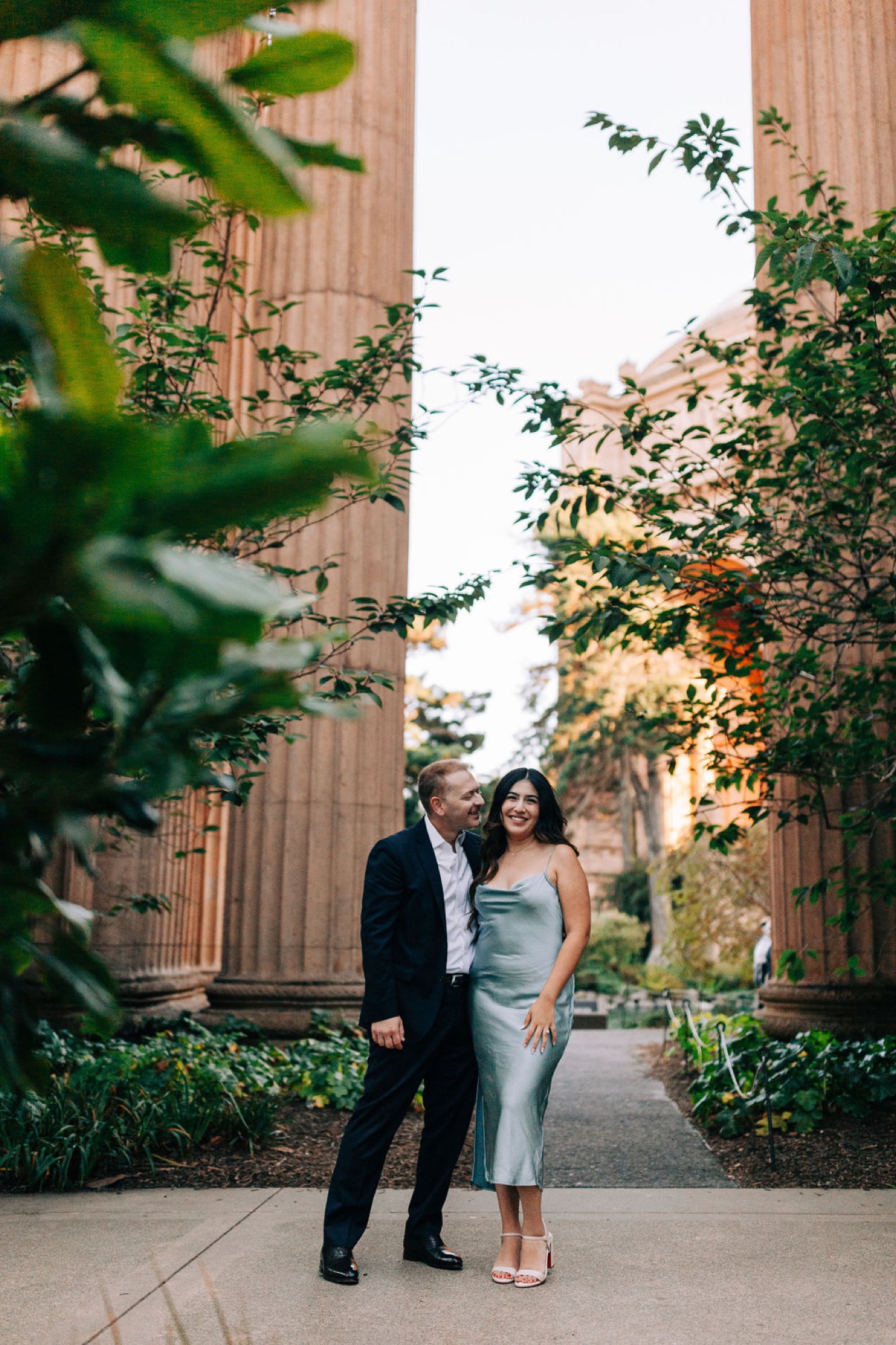 Couple stands close together between towering brick columns, soft golden afternoon light filtering through surrounding green foliage
