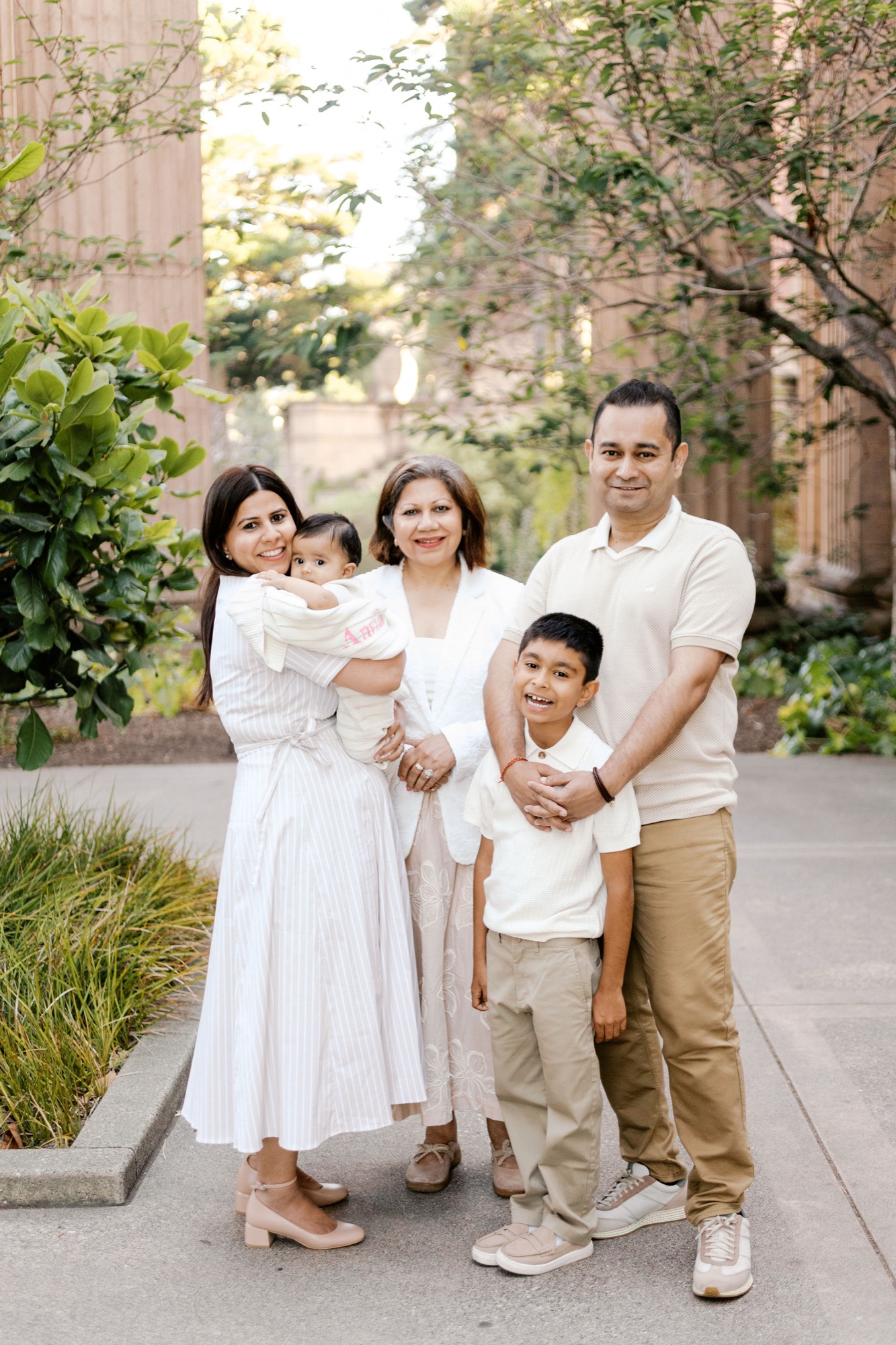 Family of five with a new baby standing among the tall columns and greenery of the Palace of Fine Arts colonnade in soft evening light