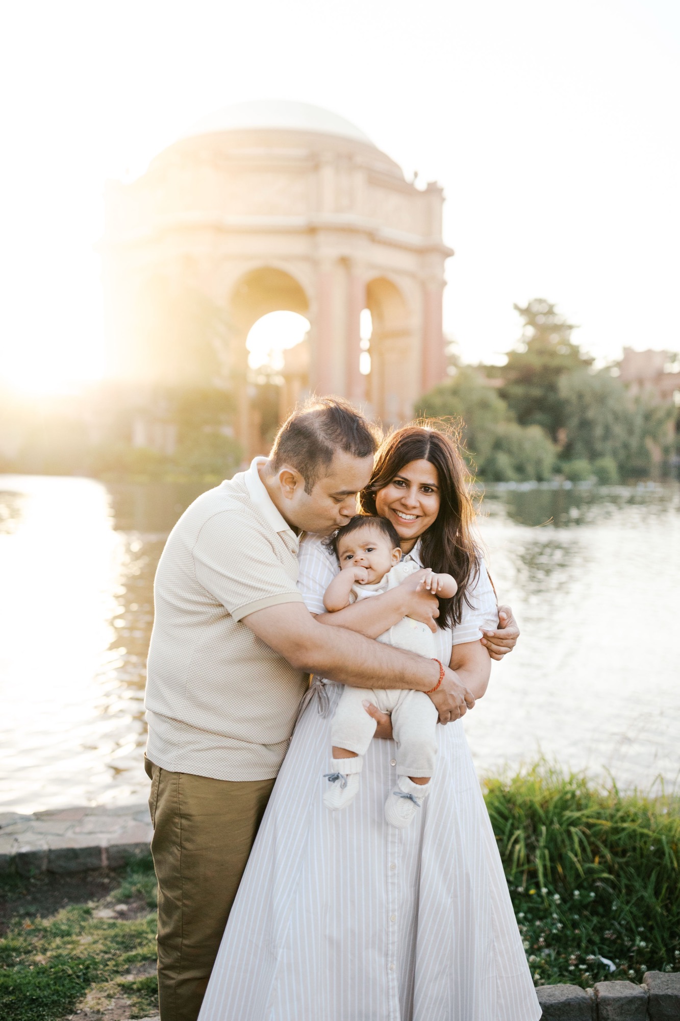 Dad kisses the top of his baby's head while mom smiles, the Palace of Fine Arts rotunda glowing across the lagoon at sunset