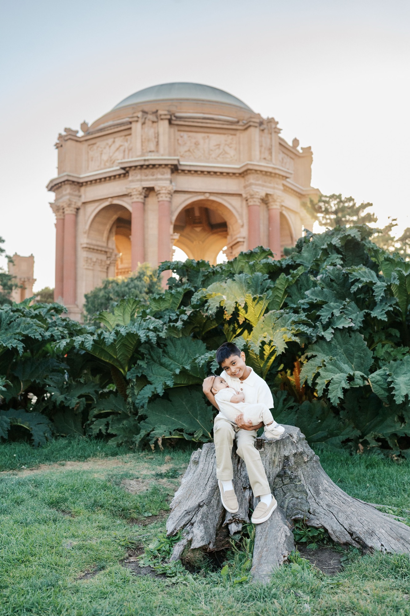 Big brother sits on a tree stump holding his baby sister, giant gunnera leaves and the Palace of Fine Arts dome behind