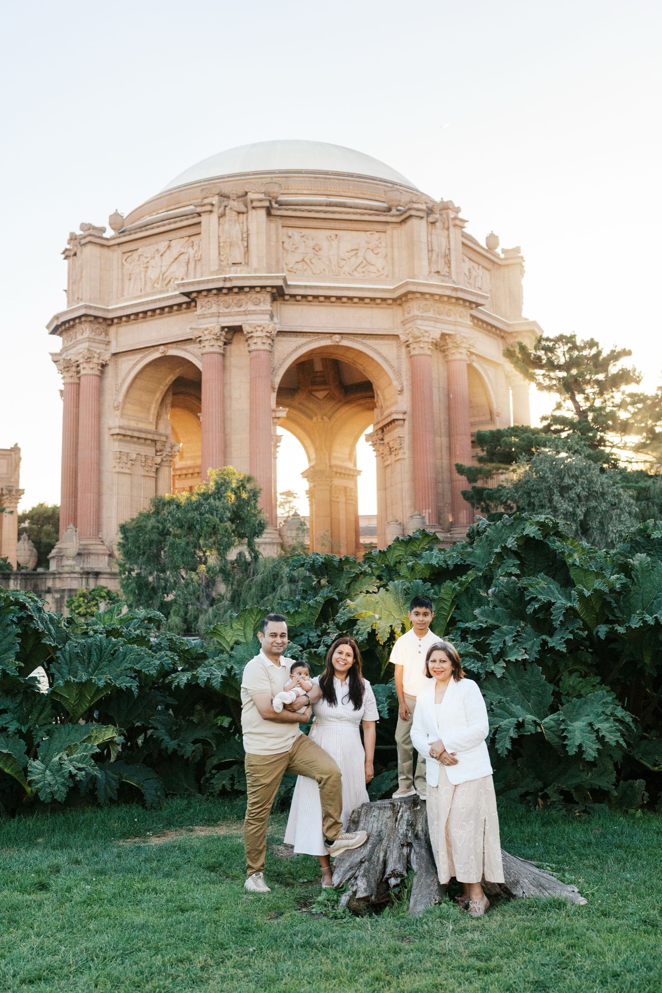 Family of five gathered around a tree stump beneath giant leaves, the Palace of Fine Arts rotunda glowing in the background