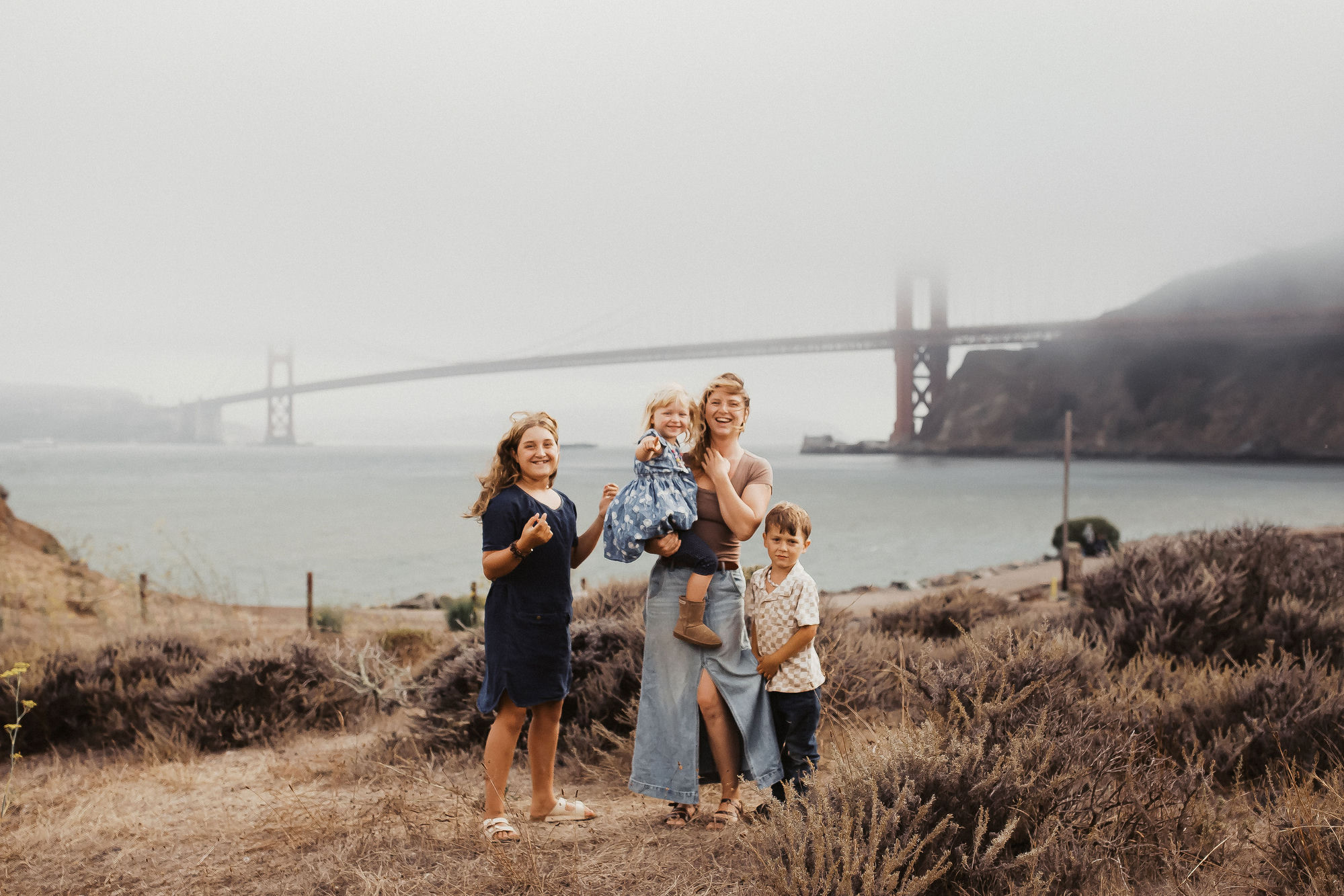 Mom holds her toddler while her two older kids gather close on a golden hillside, the Golden Gate Bridge in the fog across the bay