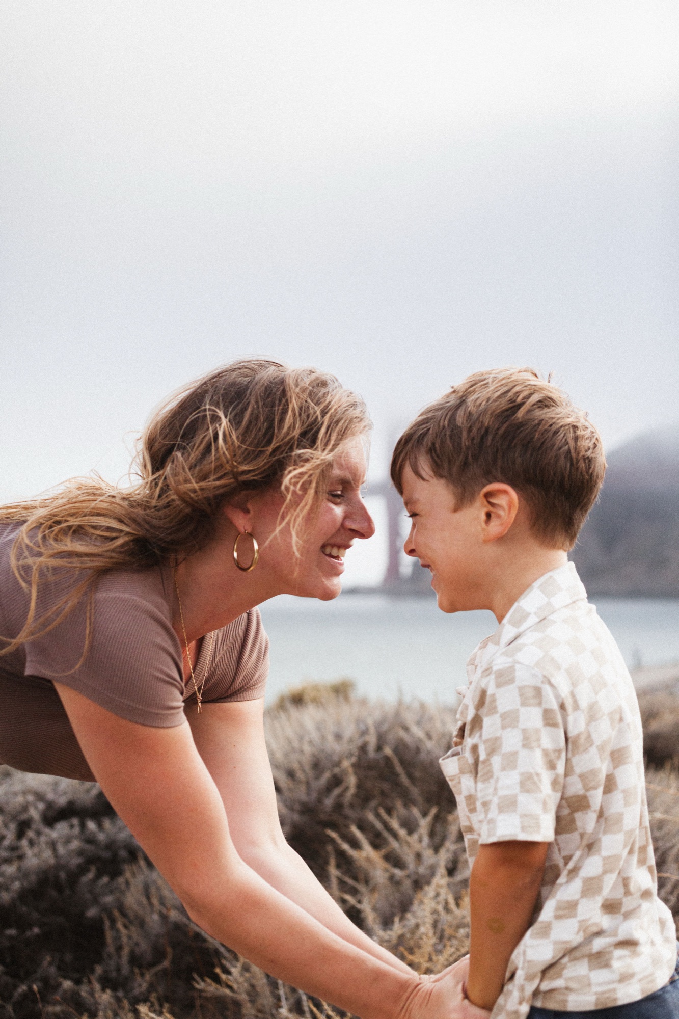 Mom leans down nose to nose with her son, laughing, the bridge tower soft in the fog behind