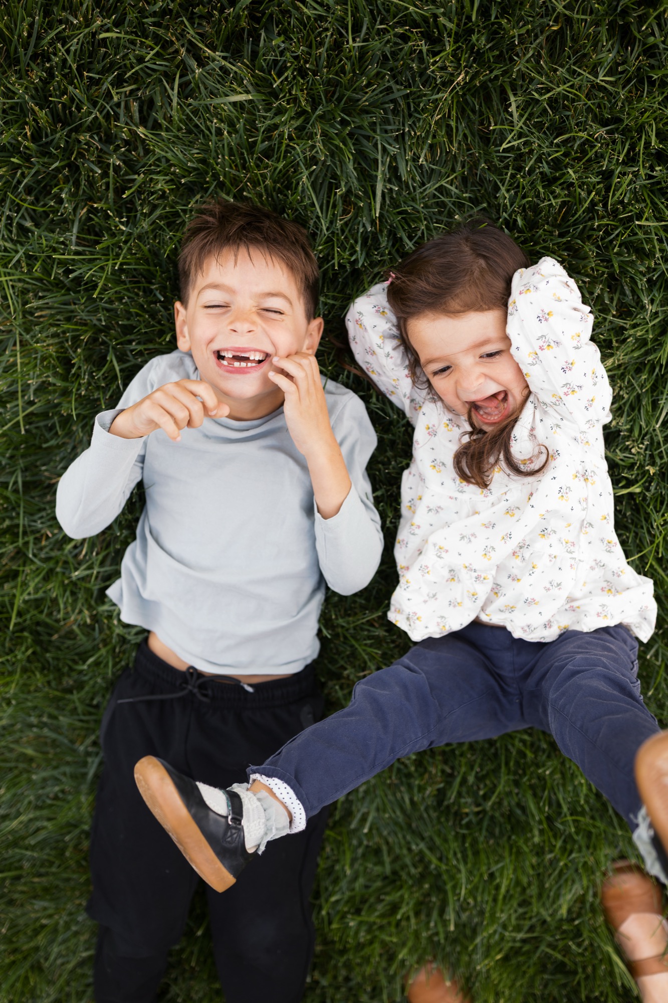 Two children lying on vibrant grass grin widely at the camera, their joyful expressions and relaxed poses capturing carefree childhood moments