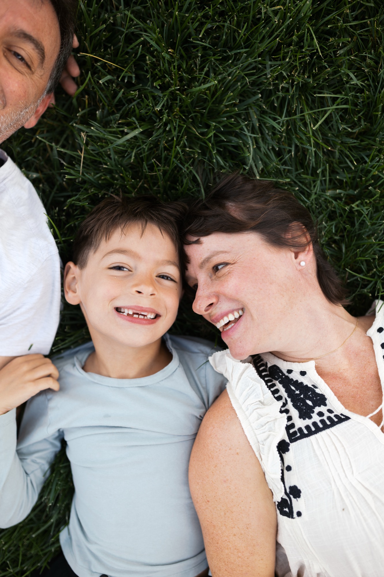 Three family members lie on grass smiling at camera, caught in a candid overhead moment with natural daylight