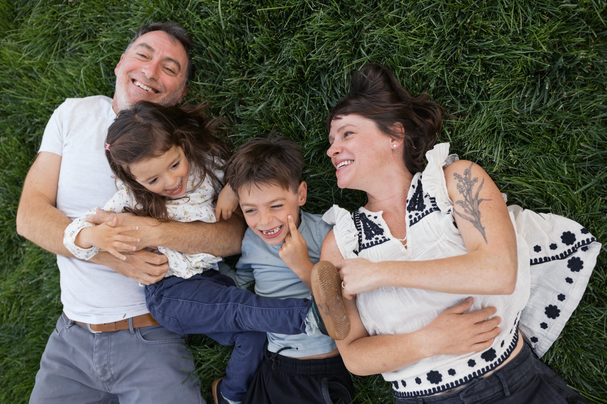 Family of four lying on grass, laughing together with genuine joy and physical closeness in bright daylight