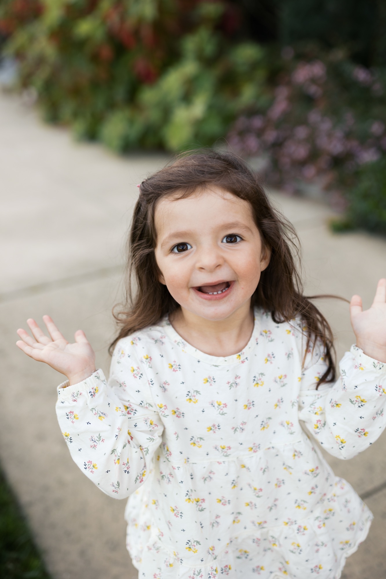 Young girl with dark hair beams at camera with both hands raised in joyful gesture, wearing cream floral print shirt