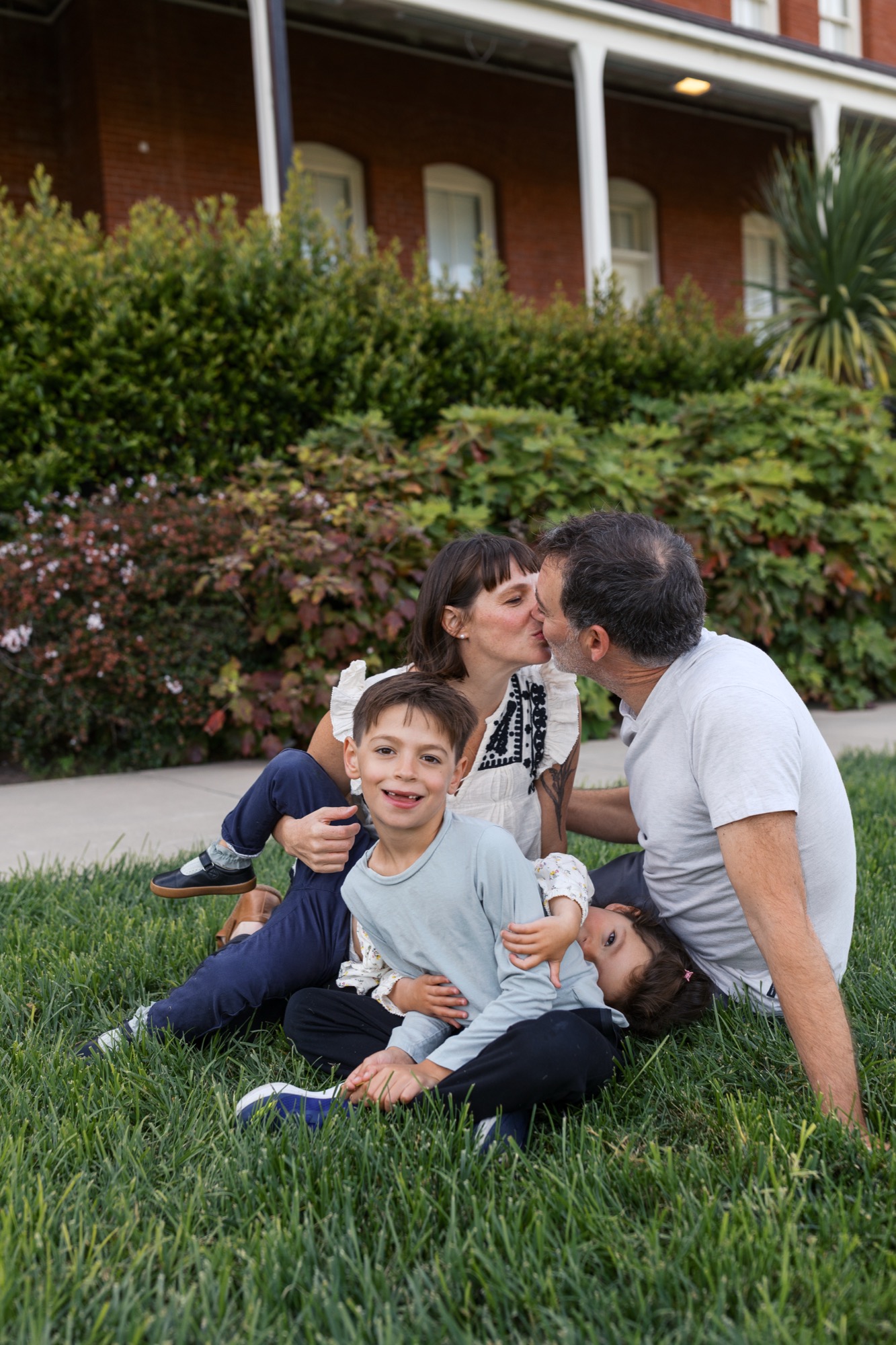 A parent and two young children sit together on grass, sharing an affectionate moment with the brick house and manicured garden behind them