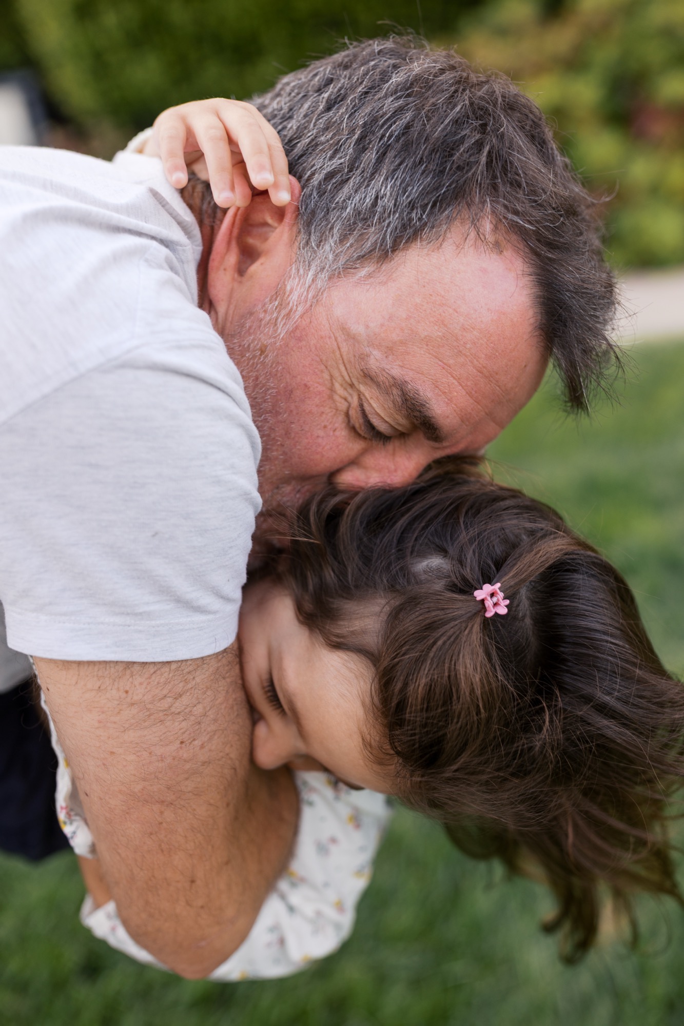 A father tenderly holds his young daughter upside down on a sunny lawn, both smiling with joy and affection