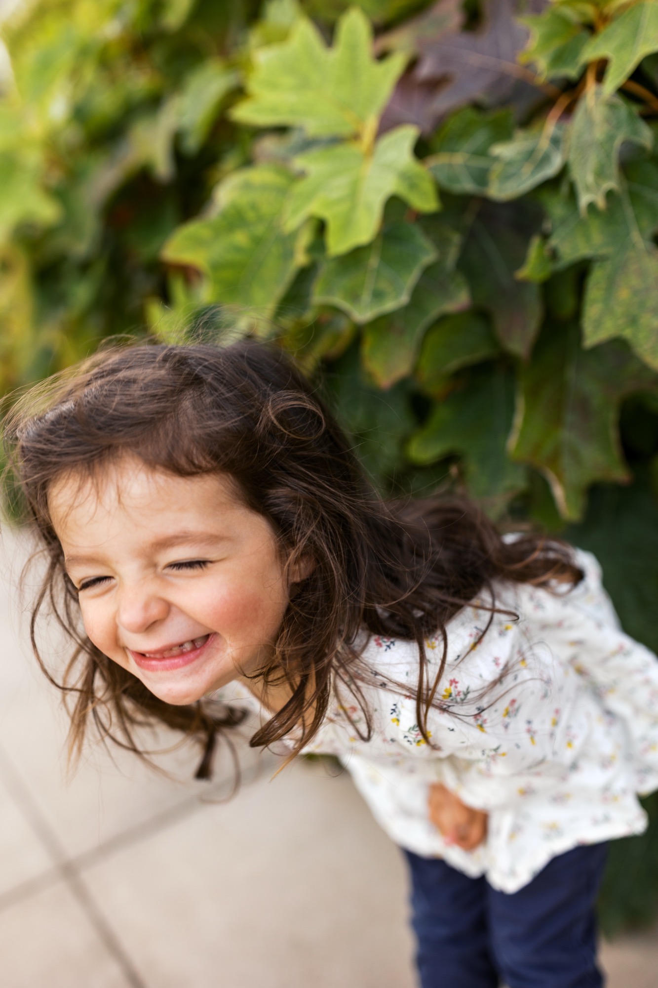 Young girl with dark wavy hair smiles warmly while standing beneath lush green ivy, wearing a white lace top and navy pants