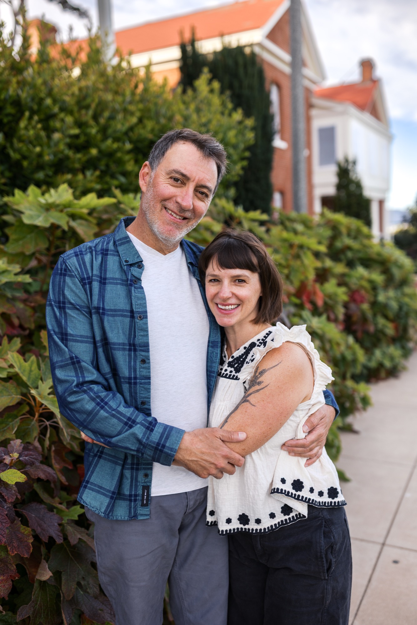 A smiling couple embraces outdoors with ivy-covered buildings and manicured hedges creating a lush backdrop in soft daylight
