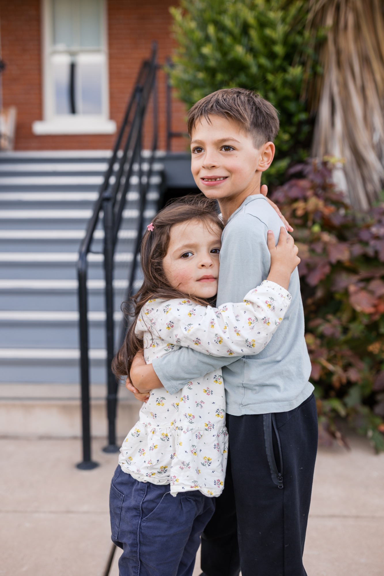 Two children embrace outdoors with genuine smiles, the boy's arm around the girl, brick building and manicured grounds behind them