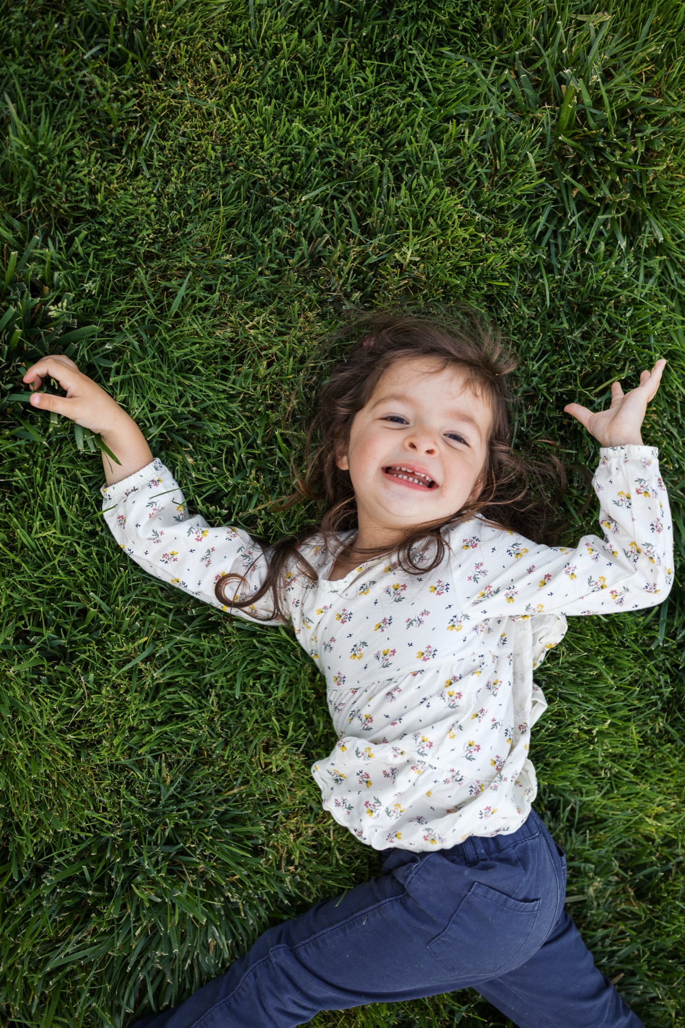 Young girl lies on vibrant grass with arms raised joyfully, wearing a patterned long-sleeve shirt and navy pants, beaming at the camera