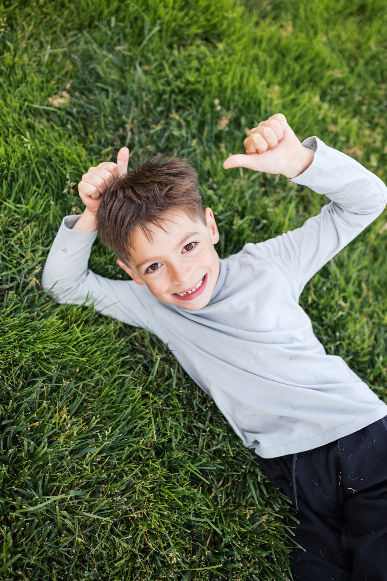 Young boy lies on vibrant green grass, arms raised overhead with genuine smile, wearing light gray long-sleeve shirt