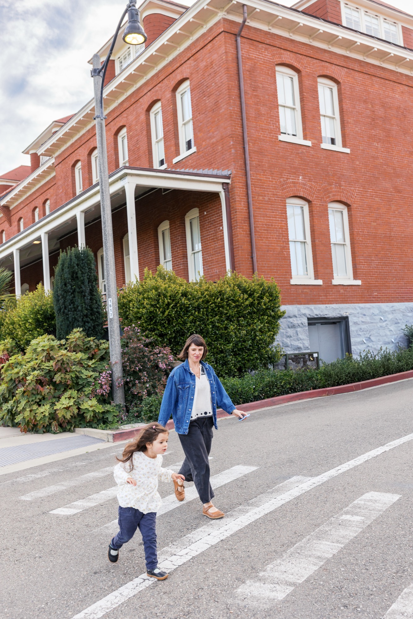 An adult and child walk across a crosswalk toward a historic red brick building on a bright, partly cloudy day