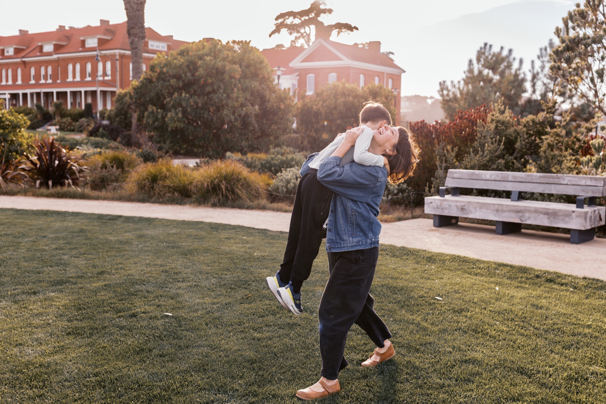 A parent lifts their child high in the air on an open lawn, both laughing with pure joy as golden afternoon light bathes the historic red brick building behind them