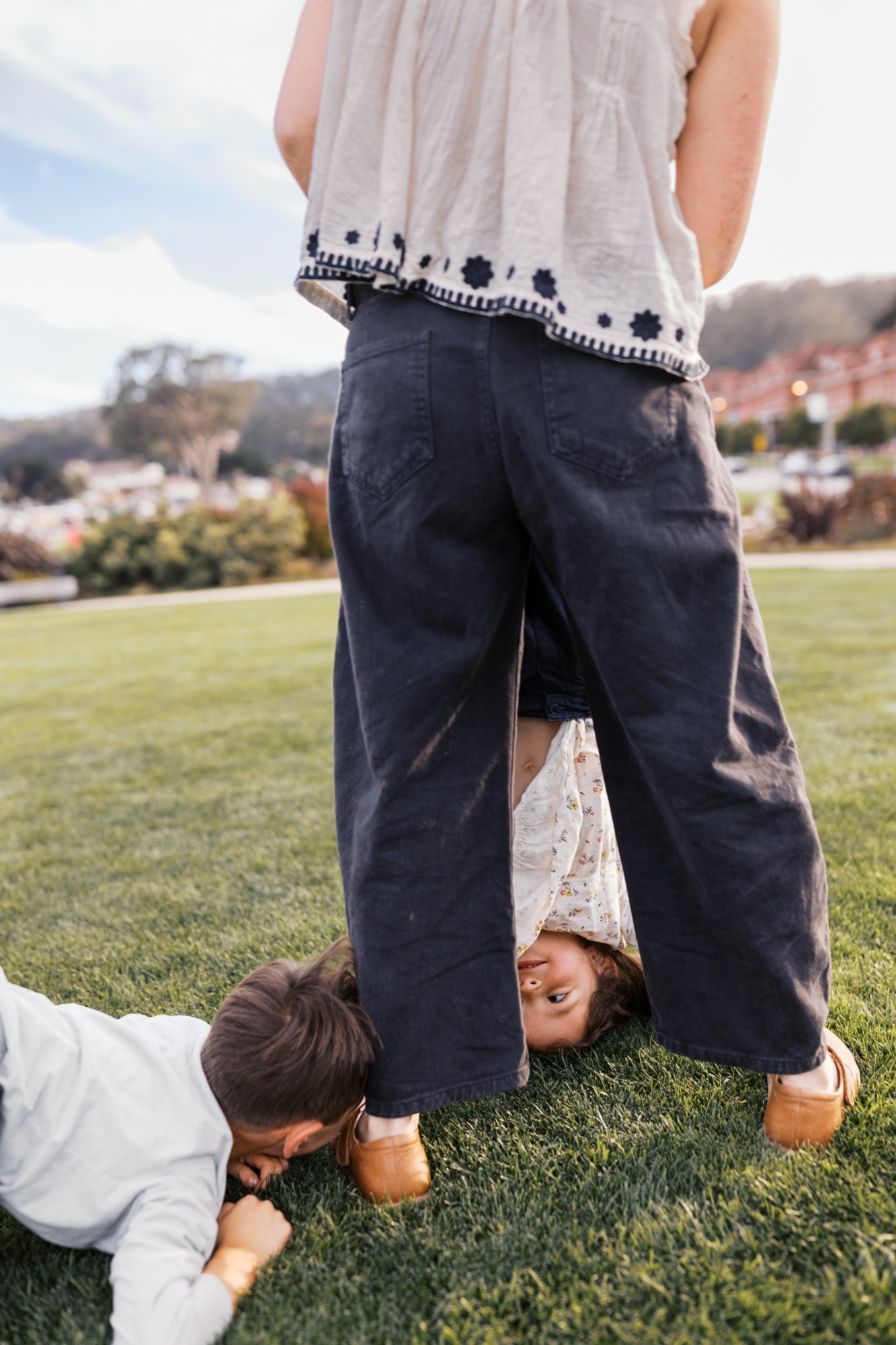 A child lies playfully on grass between an adult's legs, their head tilted back in joyful abandon on a sunny afternoon