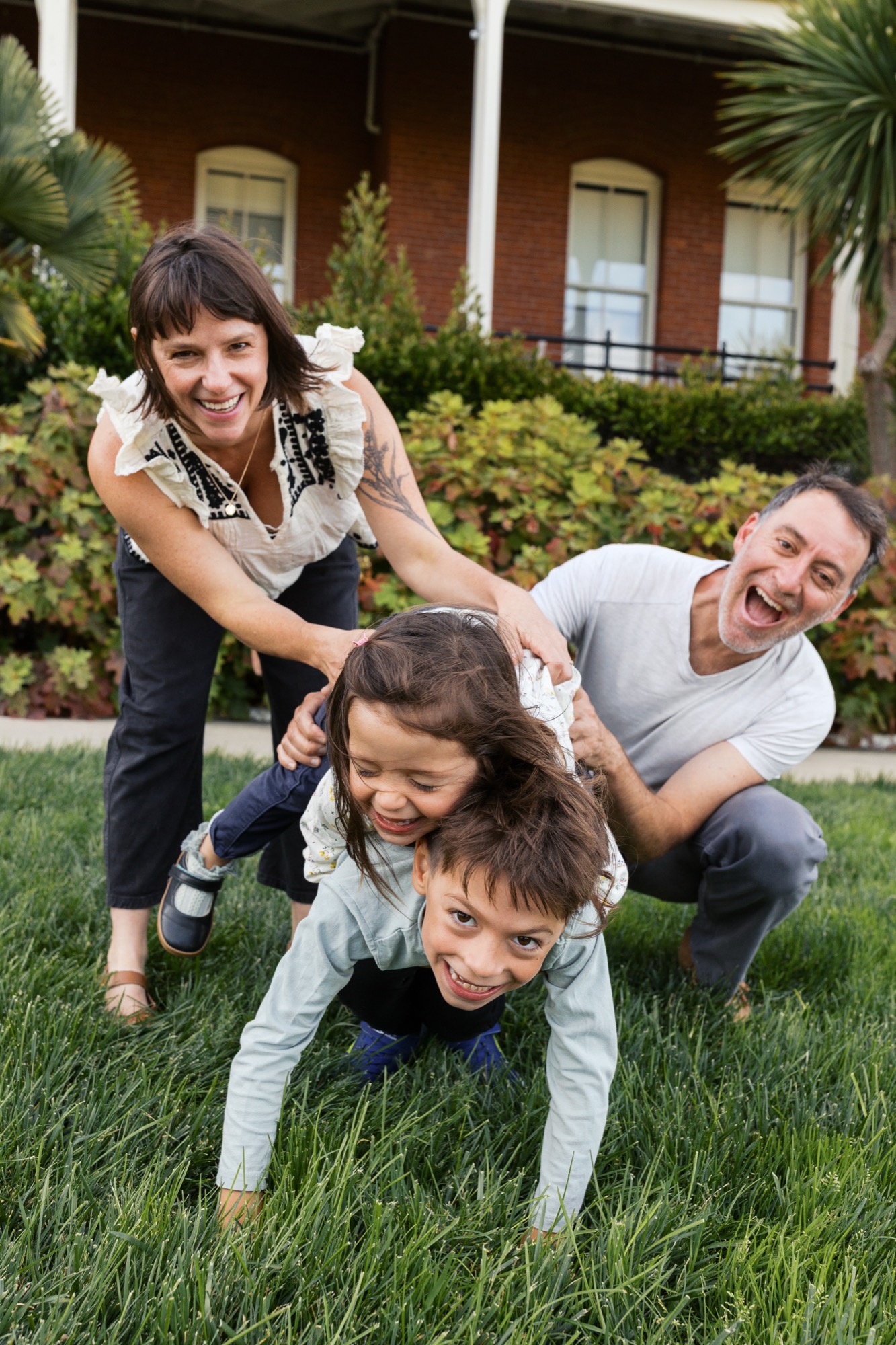 Two adults crouch playfully behind two young children on manicured grass, all beaming with genuine joy against a brick colonial home