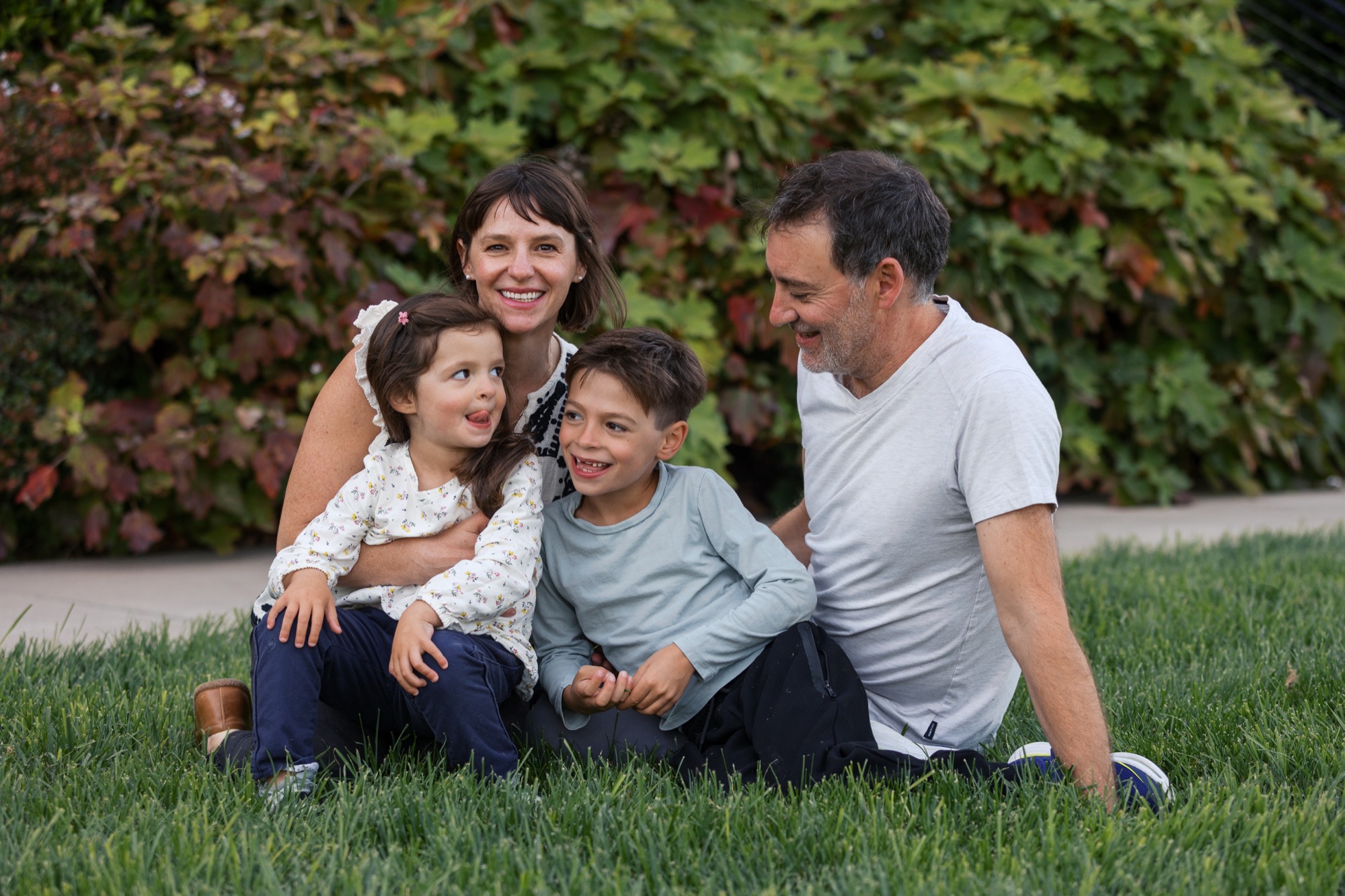 A family of four sits together on grass, smiling warmly at the camera with lush green foliage creating a natural backdrop behind them
