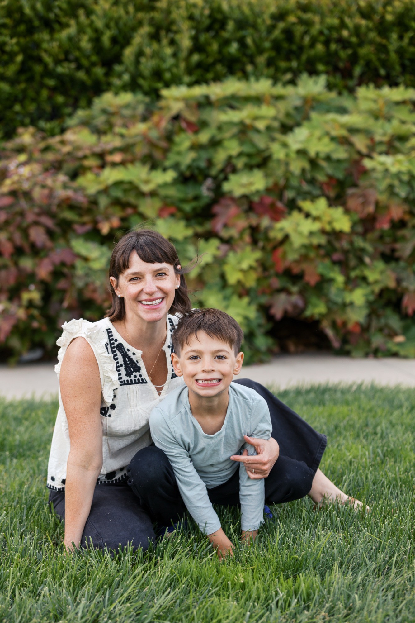 A smiling woman and young boy kneel together on manicured grass, sharing genuine joy with warm afternoon light illuminating their faces