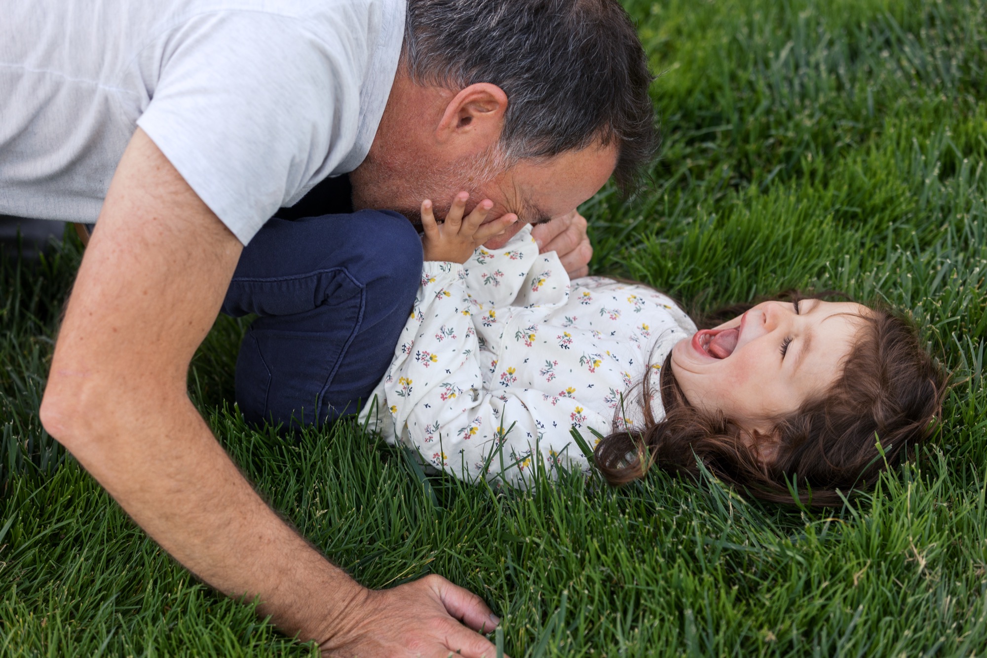 A man and young child lie on lush grass, their faces close together in a playful moment of connection and joy