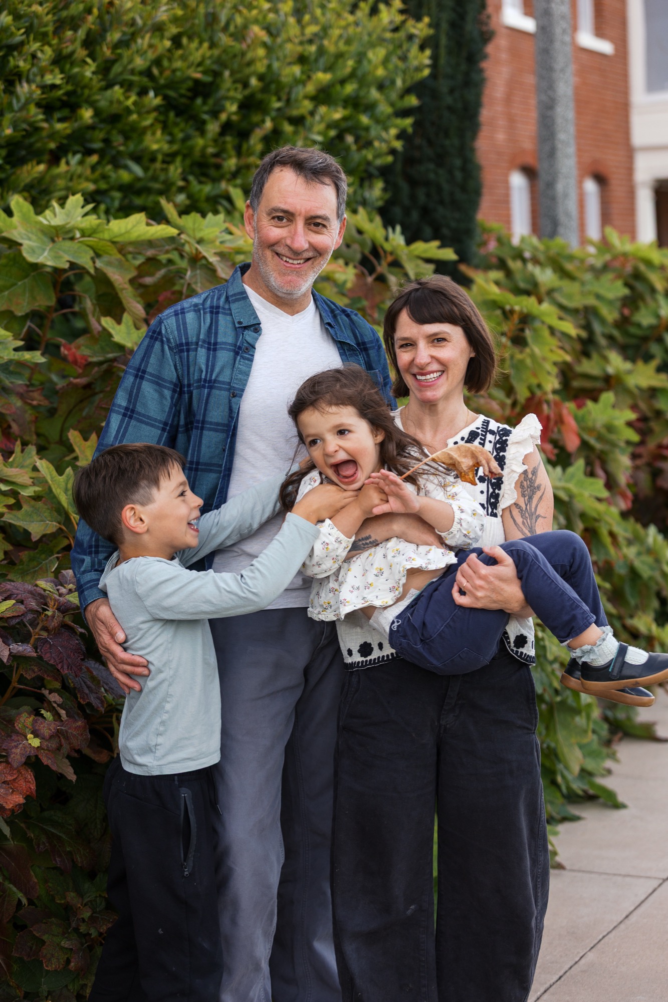 A smiling family of four stands together against lush green foliage, the father in a blue plaid shirt and mother in patterned top flanking two young children