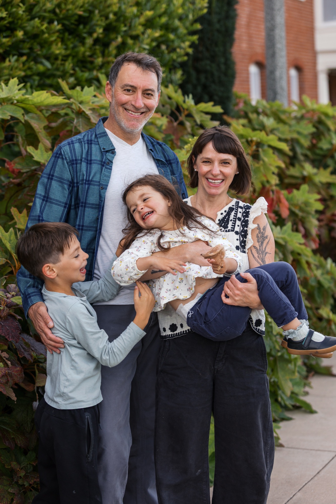 Family of four smiling warmly outdoors, father in plaid shirt holding young daughter while mother and son stand close beside them against green foliage