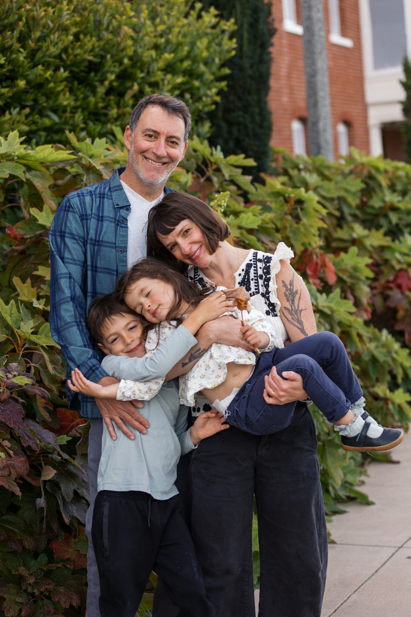 Father in plaid shirt holds three children close against his chest, all smiling warmly at camera with ivy-covered brick building behind