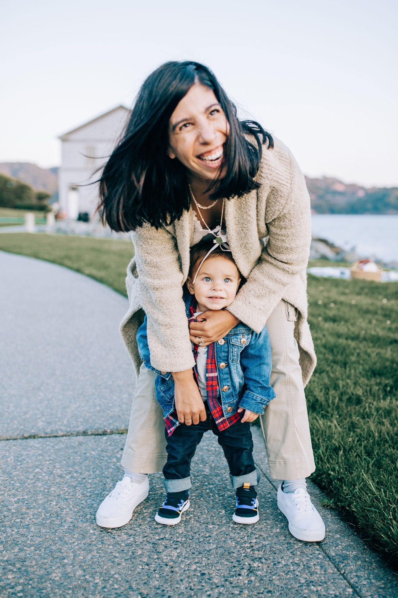 A woman in a beige cardigan bends forward to embrace her toddler on a paved path, with a calm bay and hillside homes visible behind them