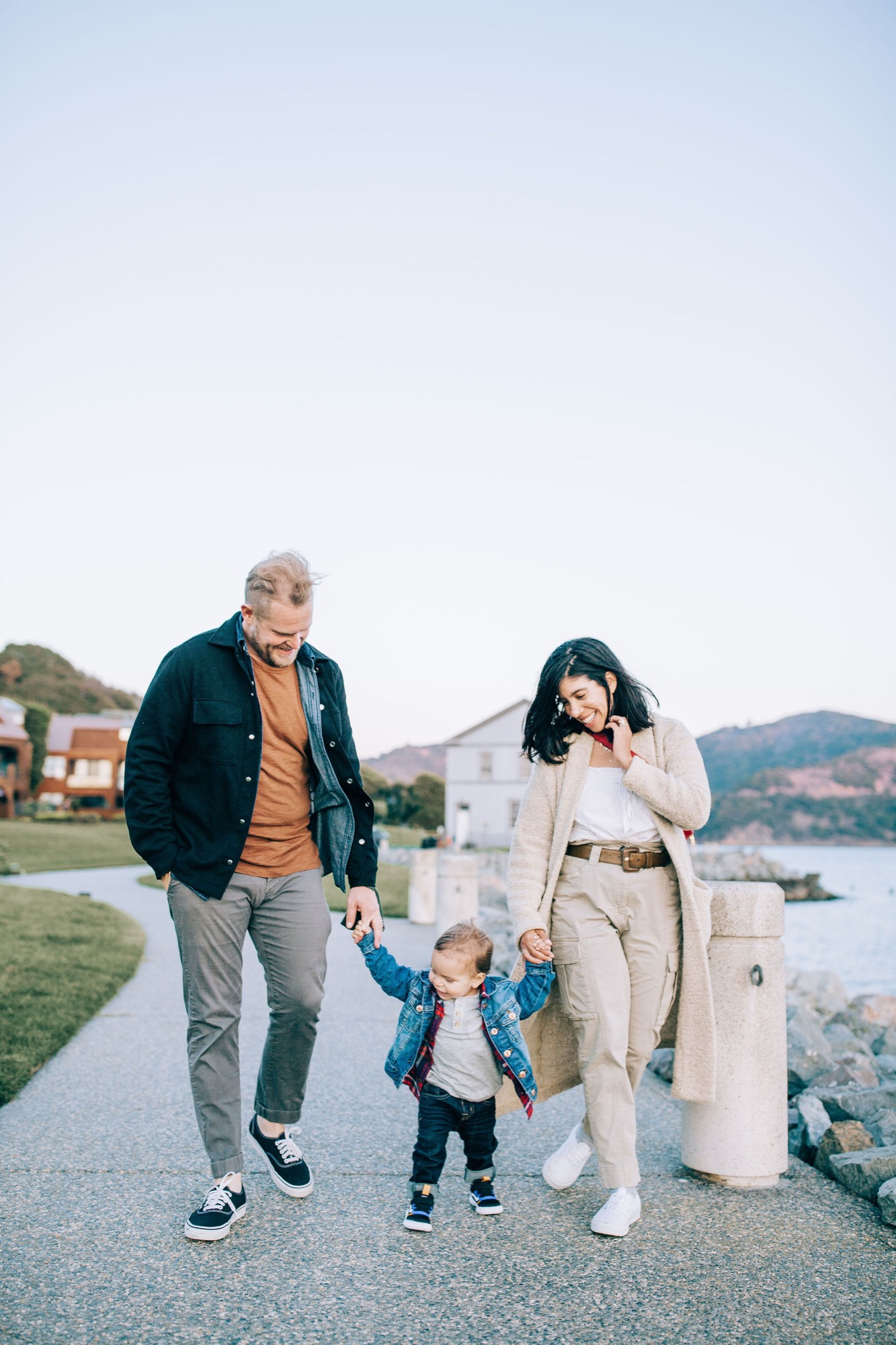 A young family walks along a waterfront path with their toddler between them, mountains and bay visible behind under overcast skies