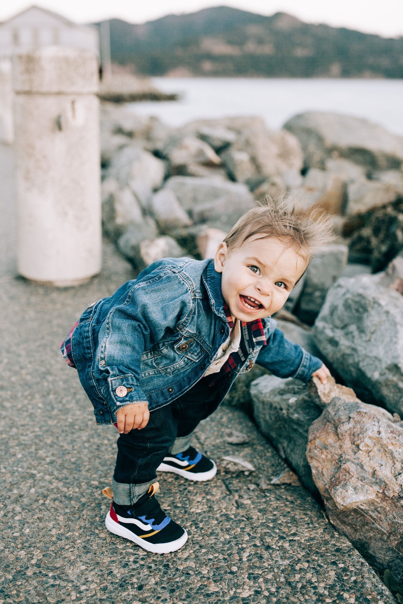 Toddler in denim jacket grins widely while crouching among rocky shoreline boulders with calm bay waters behind