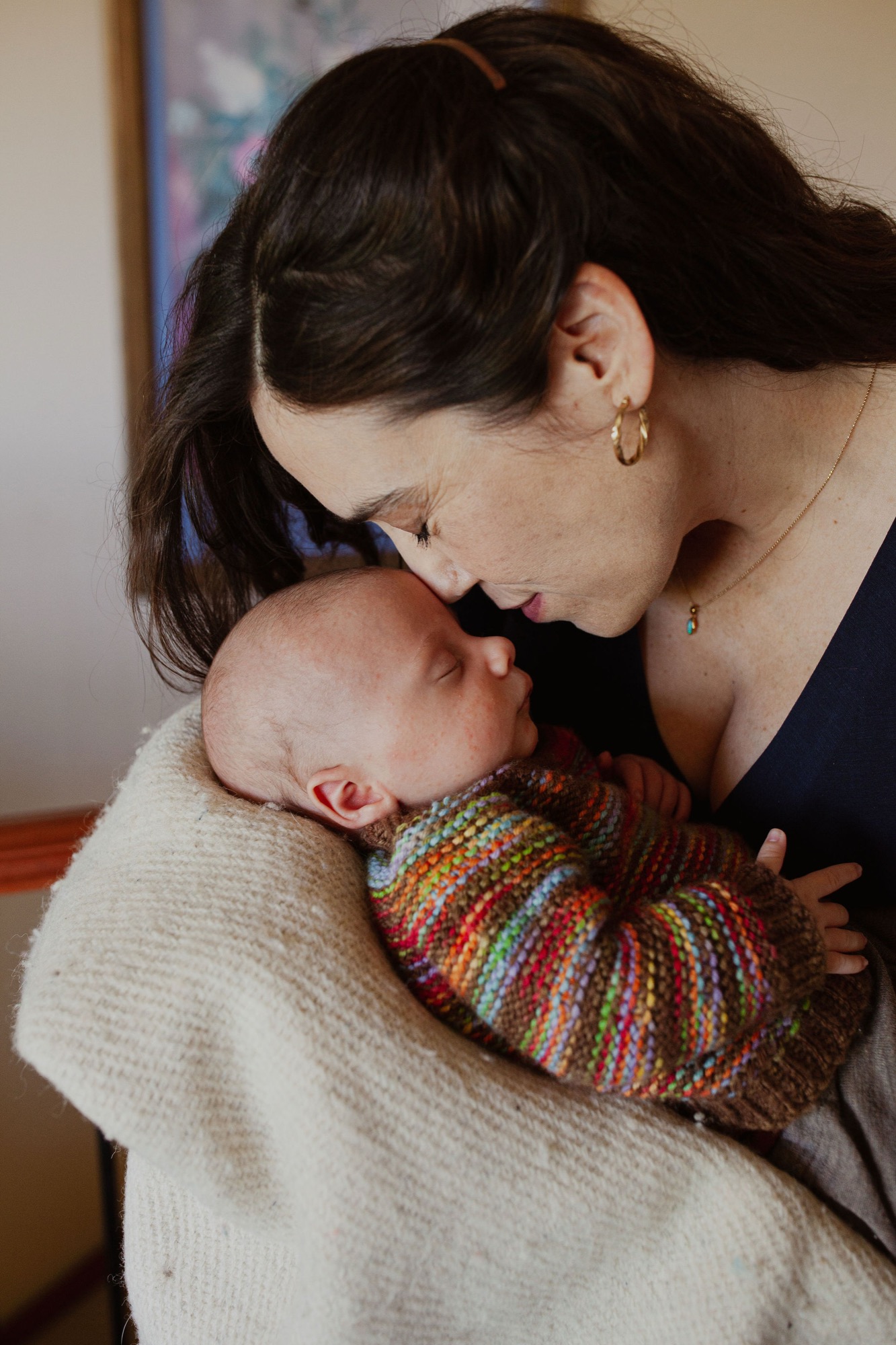 Mother gazes tenderly at newborn wrapped in colorful striped blanket, soft natural light illuminating their intimate indoor moment