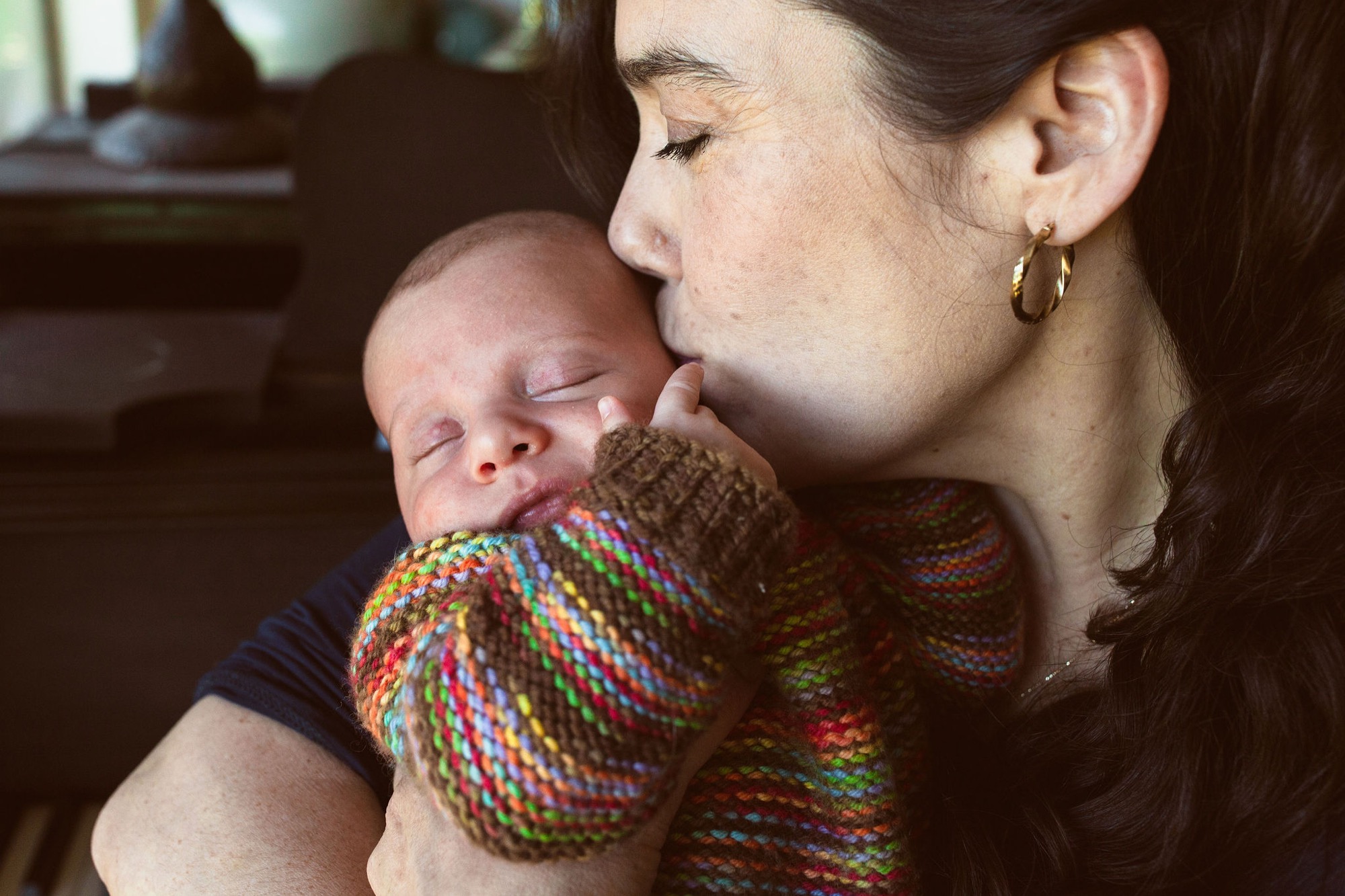 Mother kisses her newborn's forehead while cradling him in a colorful knit blanket, eyes closed in tender affection