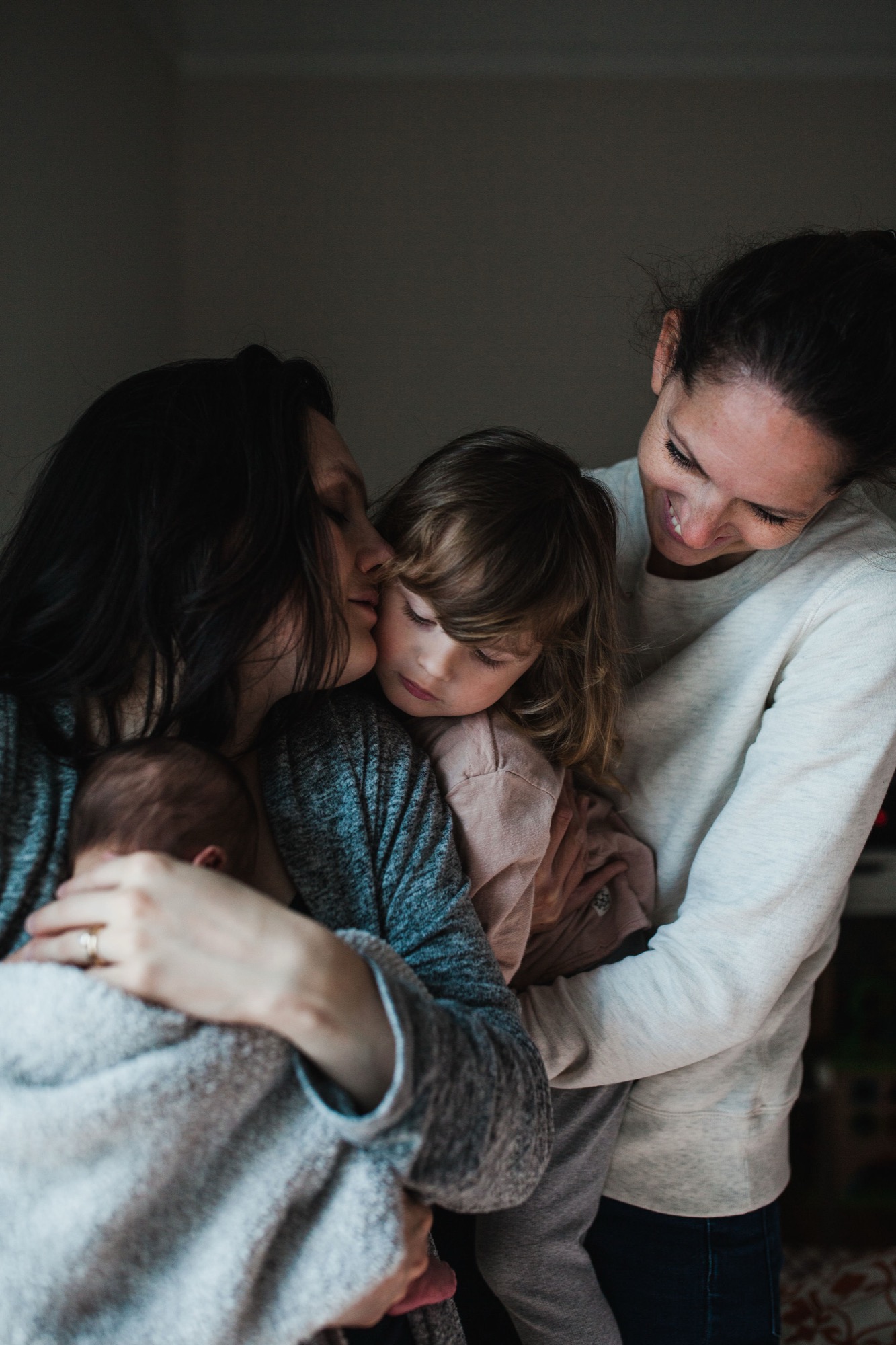 Two adults embrace a young child between them in warm indoor light, their faces close together in genuine affection and joy