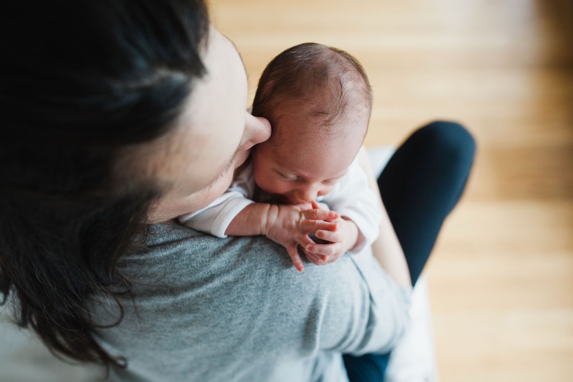 A parent in a gray shirt cradles their newborn, whose tiny hand reaches upward while resting against their shoulder in warm natural light