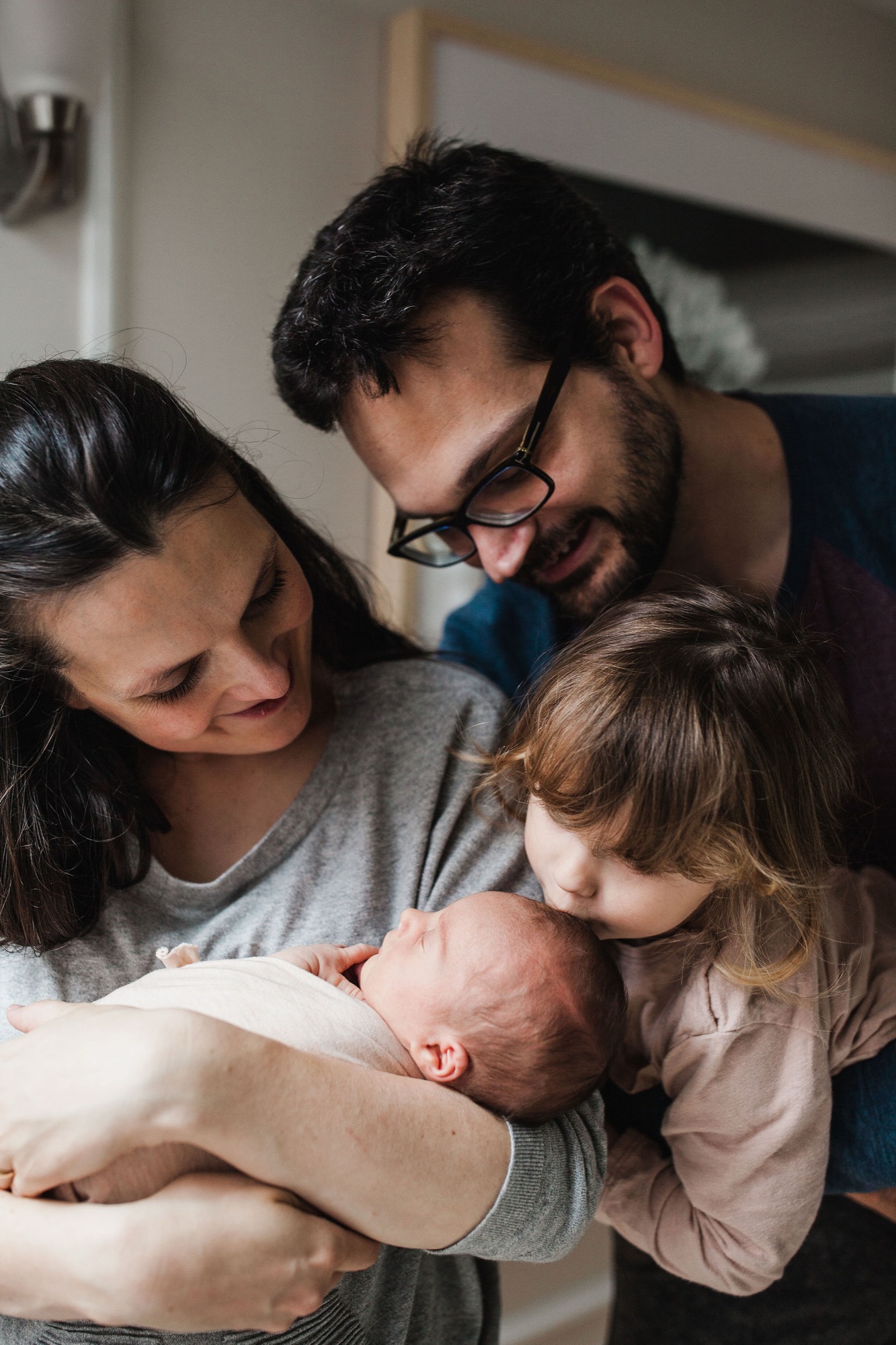 A bearded father in glasses leans tenderly over his sleeping newborn while the mother and older daughter gaze down with gentle affection in warm indoor light