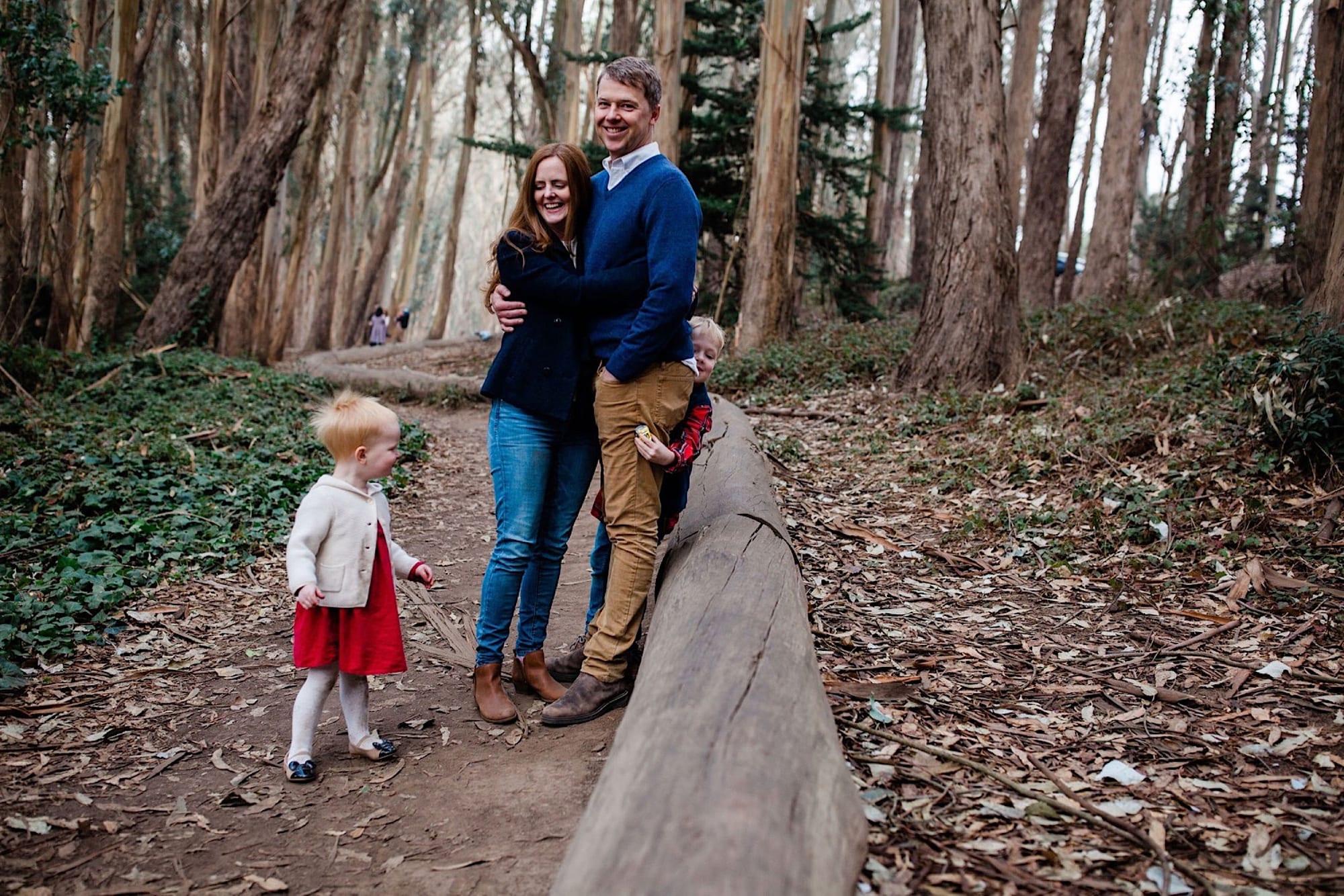 Parents holding each other on the eucalyptus-lined path at Lovers Lane while their toddler walks ahead in a red dress