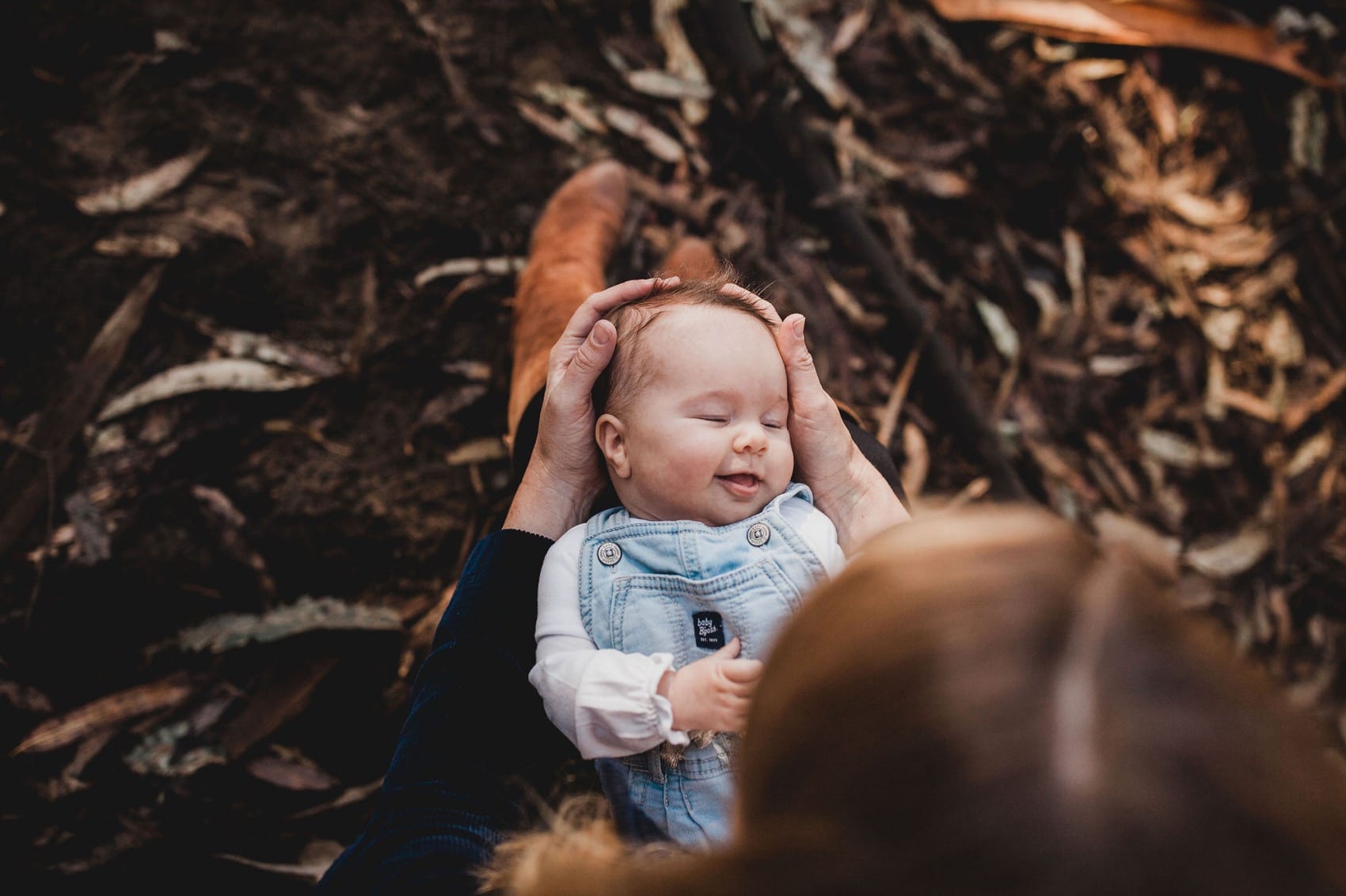 Baby in denim overalls held up in a parent's hands against the bark and leaf litter of the Presidio forest floor